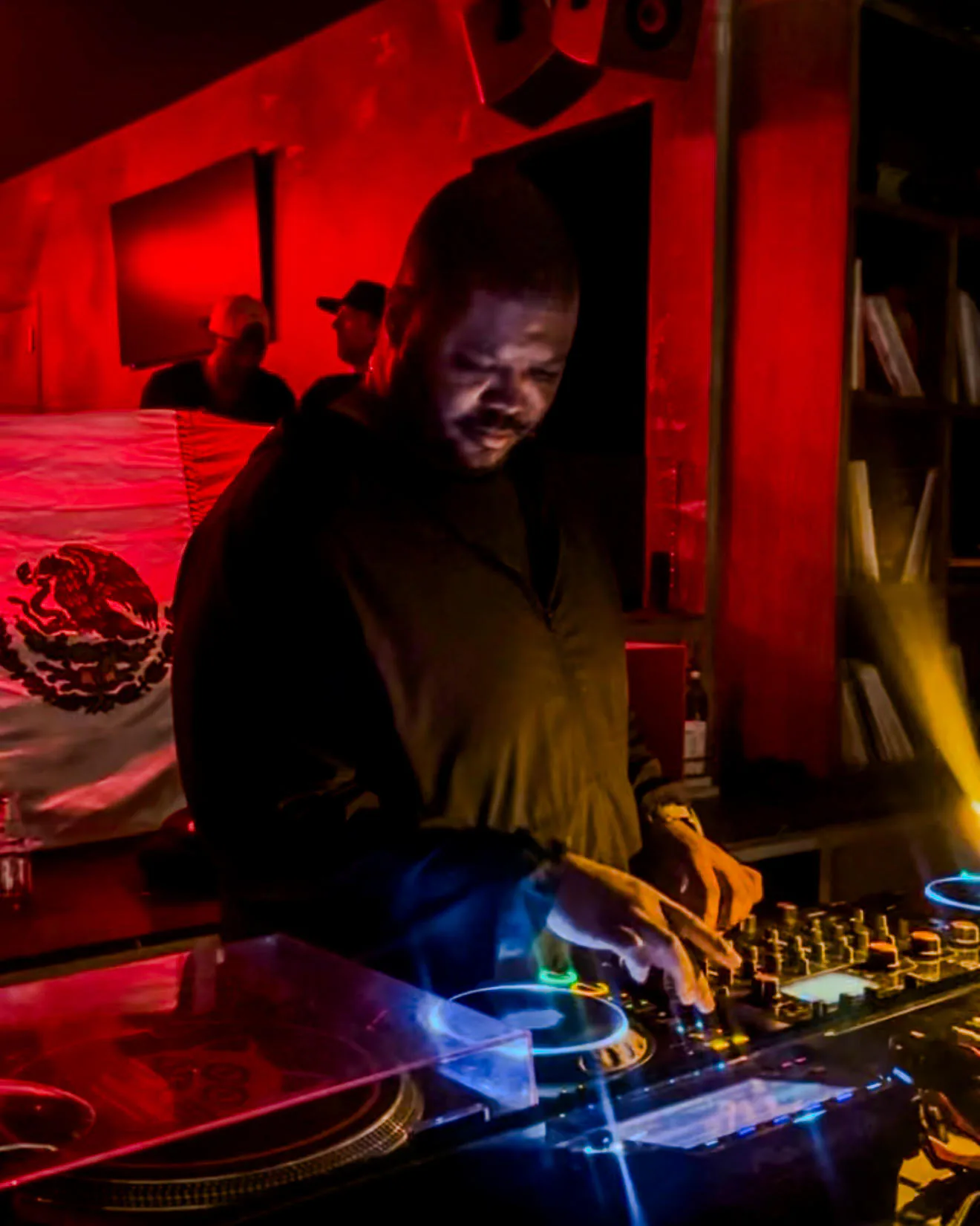 Denzel playing a club night under red light, leaning over a turntable and mixer with one hand on the controls. A Mexican flag hangs on the wall behind him and two people stand at the back of the room.