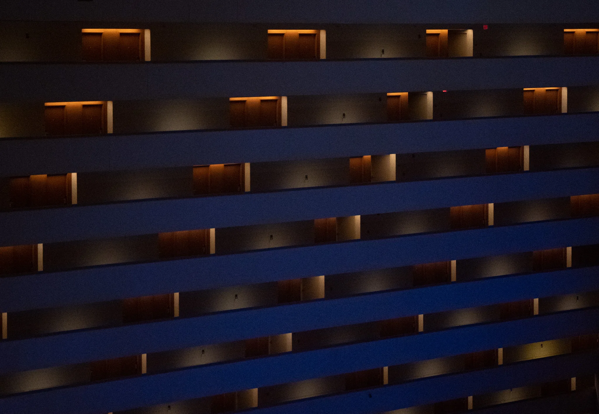 Stacked tiers of hotel balconies at night, receding diagonally across the frame. Warm lamplight picks out each doorway against the deep blue of the unlit concrete.