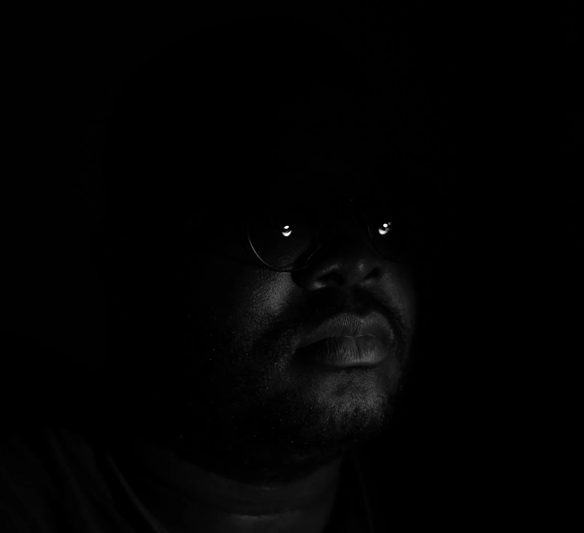 A very low-key portrait: a face emerging from near-total black, lit only enough to describe the nose, mouth and jaw. Two small highlights catch in the lenses of round glasses.
