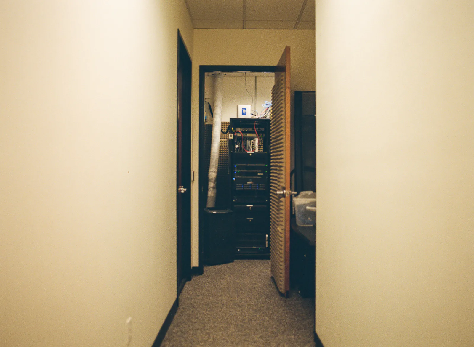 A corridor ending at a propped-open door onto a small server room: one rack of switches and servers standing under its own status lights, acoustic foam on the wall beside it and a flexible duct running up into the ceiling.